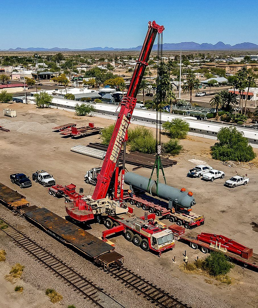 A large red crane lifting a cylindrical tank near railway tracks in a residential area.