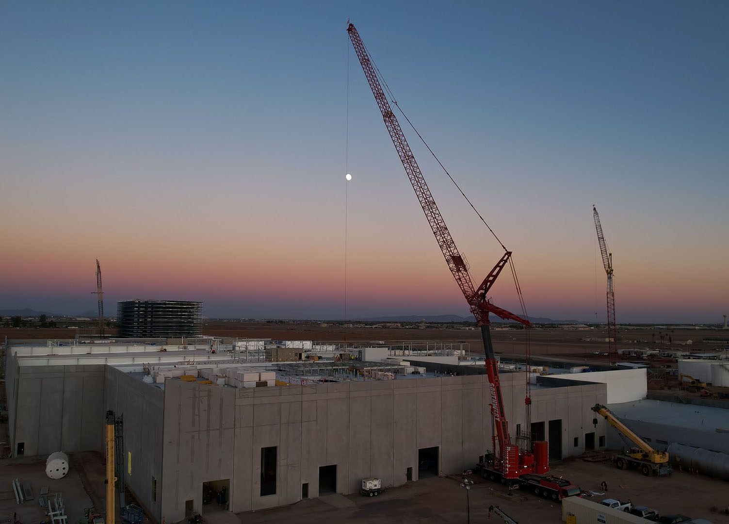 Cranes at a construction site during sunset with a clear sky and moon visible.
