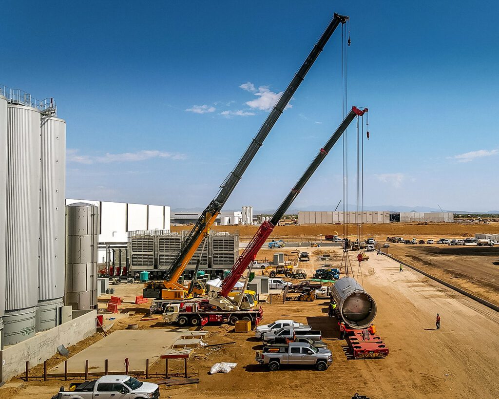 Two cranes lifting large industrial equipment at a construction site under a clear sky.