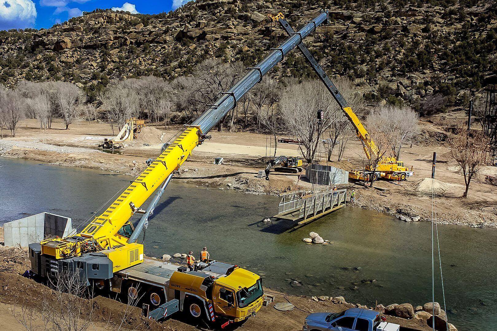 Two cranes lifting and positioning a large structure over water at a construction site.
